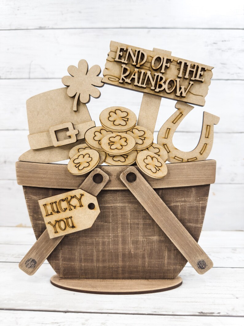 May include: Wooden craft decoration featuring a basket filled with gold coins, a shamrock, a leprechaun hat, a horseshoe, and a sign that reads "End of the Rainbow". The basket has a tag that says "Lucky You".