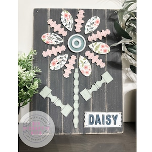 May include: A wooden sign with a daisy design made from various wooden pieces. The daisy has a pink and white floral pattern and a teal center. The sign is painted black and has a white distressed finish. The sign also has a metal sign with the word "DAISY" in white letters on a blue background.