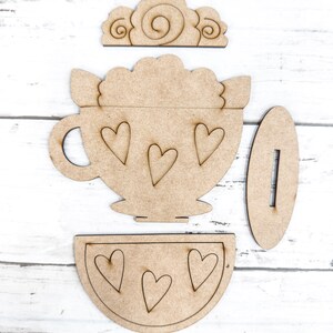 Op de afbeelding: Een houten knutselset om een ​​theekopje met een schoteltje en een deksel te maken. Het theekopje heeft een hartvormig ontwerp en het deksel heeft een swirl-ontwerp.