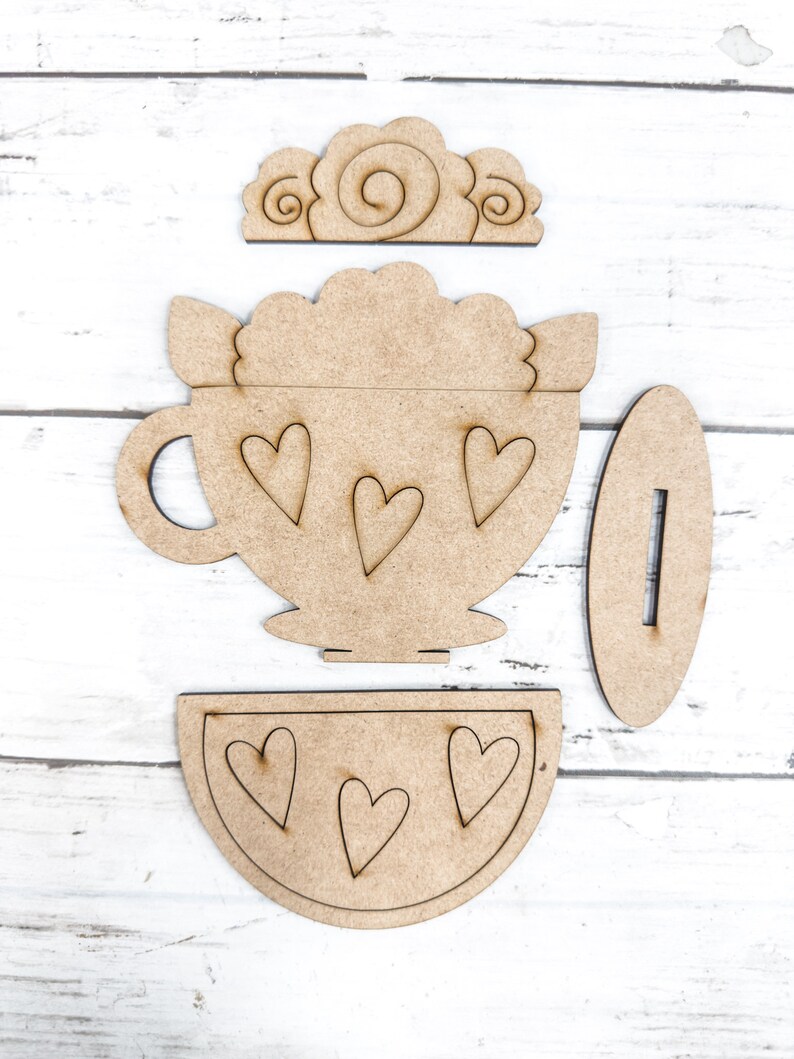 Op de afbeelding: Een houten knutselset om een ​​theekopje met een schoteltje en een deksel te maken. Het theekopje heeft een hartvormig ontwerp en het deksel heeft een swirl-ontwerp.