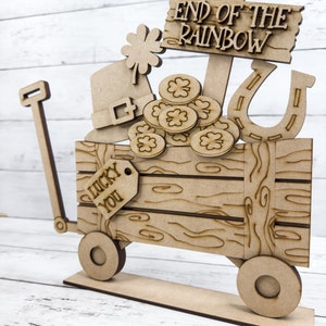 May include: Wooden cutout of a wagon with a sign that reads "End of the Rainbow". The wagon is filled with shamrocks, gold coins, a leprechaun hat, and a horseshoe. A tag on the side of the wagon reads "Lucky You".