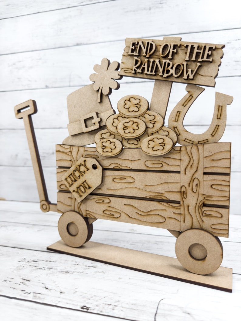 May include: Wooden cutout of a wagon with a sign that reads "End of the Rainbow". The wagon is filled with shamrocks, gold coins, a leprechaun hat, and a horseshoe. A tag on the side of the wagon reads "Lucky You".