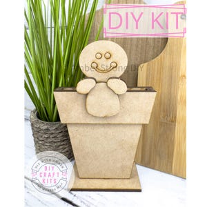 May include: DIY craft kit featuring a gingerbread man peeking over the top of a wooden flower pot. The kit includes all the pieces needed to assemble the planter. The kit is labeled "DIY Craft Kits" with a small flower graphic.
