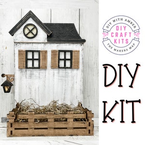 May include: DIY craft kit featuring a miniature white house with black shutters and a brown roof. The house is sitting on a wooden base with a brown fence and straw inside. The text "DIY WITH AMBER DIY CRAFT KITS THE MAKERS MAP" is on a pink circle with a white border. The text "DIY KIT" is in black with a pink outline.