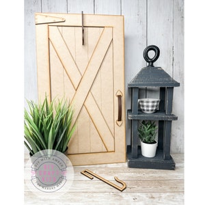 May include: A black wooden lantern with three shelves, a white and black checkered bowl, a small potted succulent, and a wooden door with a cross design. The lantern is sitting on a wooden surface. The text "DIY WITH AMBER DIY CRAFT KITS THE MAKERS WAY" is on a pink and white circle.