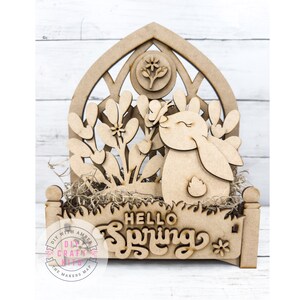 May include: Wooden cutout of a bunny rabbit sniffing a flower with the text "Hello Spring" on a wooden base. The cutout is in a light brown color and has a rustic, farmhouse style.