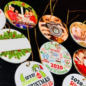 Peut inclure: Un ensemble de boules de No&euml;l rondes en c&eacute;ramique avec diff&eacute;rents motifs. Certaines boules pr&eacute;sentent des photos de famille, d'autres des motifs sur le th&egrave;me des f&ecirc;tes, et l'une d'elles a un espace blanc vide. Les boules sont d&eacute;cor&eacute;es de feuilles de houx vertes et de baies rouges.