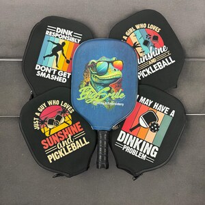 May include: Five different pickleball paddle covers with various designs. One cover features a green lizard with sunglasses and the text "Big Smile Printing & Embroidery". Another cover has a sunset scene with the text "Just a guy who loves sunshine and pickleball". A third cover has a sunset scene with the text "A girl who loves sunshine and pickleball". A fourth cover has a sunset scene with the text "Dinking Problem". The fifth cover has a sunset scene with the text "Dink Responsibly Don't Get Smashed".