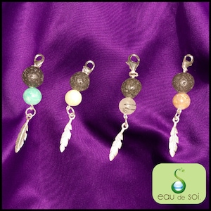 Peut inclure: Quatre colliers avec breloques en argent, des perles de lave noire, un charme en plume d'argent et des perles de pierres précieuses de différentes couleurs. Les colliers sont présentés sur un fond en tissu violet.