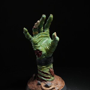 May include: A green, decaying zombie hand sculpture with a gold ring on the finger. The hand is reaching up and is attached to a base with dirt and insects.