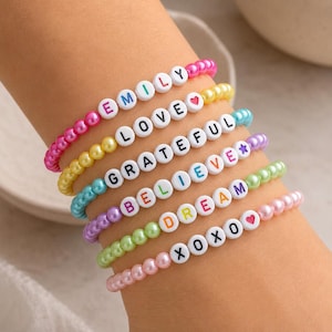 May include: Six stacked beaded bracelets featuring colourful faux pearls and white letter beads. The bracelets display the words EMILY, LOVE, GRATEFUL, BELIEVE, DREAM, and XOXO, along with a heart symbol and a star symbol in various vibrant colours.
