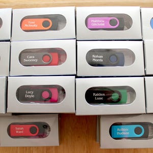 Puede incluir: Una colecci&oacute;n de memorias USB personalizadas en cajas grises individuales. Cada unidad tiene un acento de color diferente y un nombre impreso. Los colores incluyen naranja, rosa, amarillo, verde y azul.