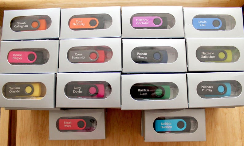 Puede incluir: Una colecci&oacute;n de memorias USB personalizadas en cajas grises individuales. Cada unidad tiene un acento de color diferente y un nombre impreso. Los colores incluyen naranja, rosa, amarillo, verde y azul.