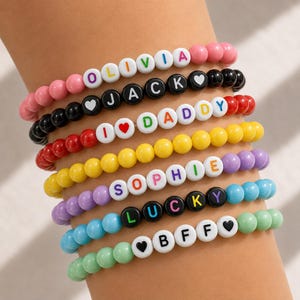 May include: A stack of eight beaded bracelets featuring colourful plastic spheres and white letter beads. The bracelets display the names OLIVIA, JACK, SOPHIE, LUCKY, and BFF, along with the phrase I LOVE DADDY, accented with heart symbols.