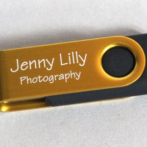 Puede incluir: Una unidad flash USB dorada y negra. La parte dorada tiene el texto "Jenny Lilly Photography" en blanco. La unidad USB tiene un dise&ntilde;o giratorio y un peque&ntilde;o lazo para sujetarla a un llavero.