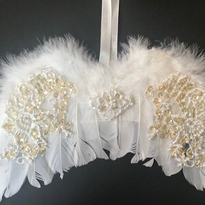 May include: White feather angel wings with gold and white beaded embellishments. The wings are attached to a white ribbon.