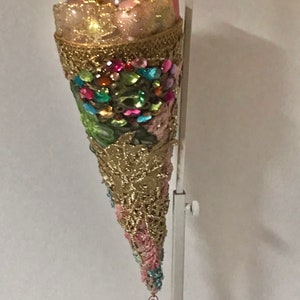 May include: A gold and pink cone-shaped Christmas ornament with a pink ribbon and a pink crystal hanging from the bottom. The cone is decorated with rhinestones and beads.