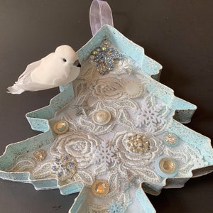 May include: A light blue Christmas tree-shaped ornament with a white feathery bird, white lace, and blue and silver embellishments.