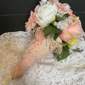 May include: A bouquet of pink and white silk flowers arranged in a cone-shaped fabric holder with lace and beaded embellishments. The bouquet is sitting on a white crocheted surface.