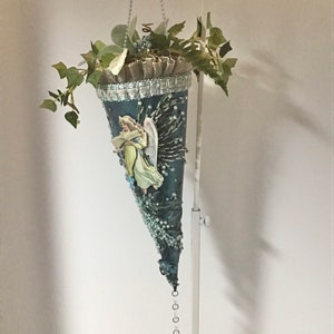 May include: A blue and white cone-shaped ornament with a beaded trim and a crystal pendant. The ornament features a vintage image of an angel with wings and a book. The ornament is decorated with greenery and is hanging from a white stand.