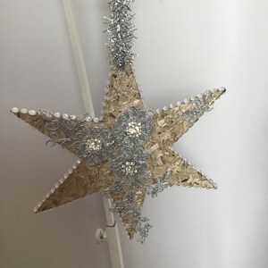 May include: A large, five-pointed star made of wood and decorated with silver glitter and rhinestones. The star is hanging from a silver string.