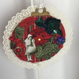 May include: A round Christmas ornament with a red and black plaid background. The ornament is decorated with a variety of embellishments, including sequins, beads, and a small picture of a girl in a white coat. The ornament is surrounded by a white lace trim.