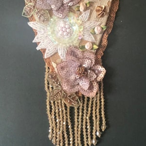 May include: A heart-shaped fabric ornament with a beaded fringe. The heart is decorated with three pink and white flowers made of sequins and beads.