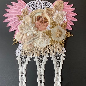 May include: A pink and white Christmas ornament with a picture of two angels, lace trim, and the text "Christmas Blessings".