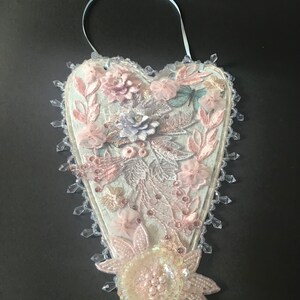 May include: A white heart-shaped ornament decorated with pink and white lace, beads, and flowers. The ornament has a light blue ribbon for hanging.
