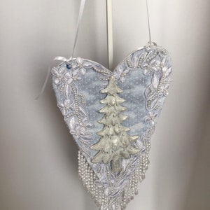 May include: A white heart-shaped ornament with a silver glitter Christmas tree design and white beaded fringe.