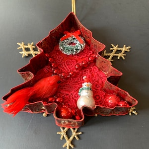 May include: A red metal Christmas tree ornament with a red cardinal, a snowman, a red and green wreath, and red beads. The ornament is decorated with red lace and gold glitter.