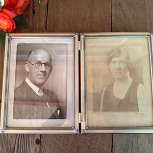 May include: A hinged double picture frame with a metallic silver finish and a beaded edge detail. The frame holds two vintage black and white portrait photographs, one of a person wearing glasses and a suit, and one of a person in a sleeveless top.