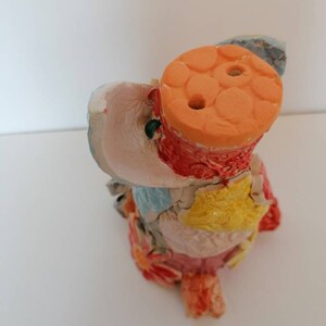 May include: A colourful ceramic sculpture of a bird with a round orange top. The bird is made of different coloured clay pieces and has a textured surface.