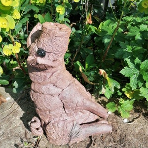 May include: A brown ceramic sculpture of a creature with a large head, two eyes, and a long tail. The sculpture is sitting on a bed of gravel.