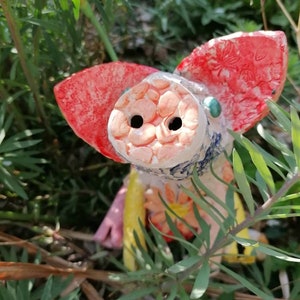 May include: A ceramic pig figurine painted in red, pink, yellow and blue. The pig has a playful expression and is partially hidden amongst green foliage.