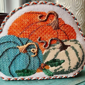 May include: A hand-stitched needlepoint pillow featuring three pumpkins in shades of orange, teal, and white. The pumpkins have detailed stitching and are surrounded by green leaves. The pillow has a white background and a decorative border.