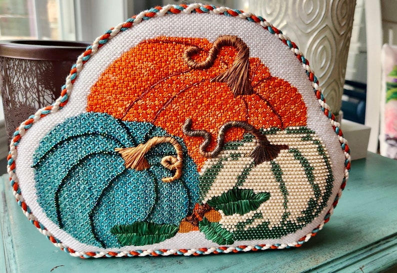 May include: A hand-stitched needlepoint pillow featuring three pumpkins in shades of orange, teal, and white. The pumpkins have detailed stitching and are surrounded by green leaves. The pillow has a white background and a decorative border.
