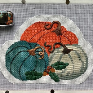 May include: A needlepoint design featuring three pumpkins in shades of orange, teal, and white. The pumpkins are arranged in a cluster with their stems intertwined. The design is set against a white background.