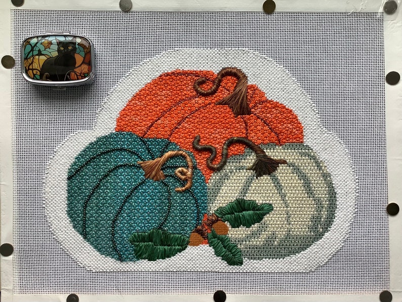 May include: A needlepoint design featuring three pumpkins in shades of orange, teal, and white. The pumpkins are arranged in a cluster with their stems intertwined. The design is set against a white background.