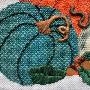 May include: Close-up of a teal blue pumpkin with a brown stem and green leaves. The pumpkin is embroidered with beads and the stem is embroidered with brown thread.