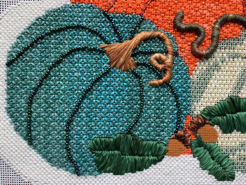 May include: Close-up of a teal blue pumpkin with a brown stem and green leaves. The pumpkin is embroidered with beads and the stem is embroidered with brown thread.
