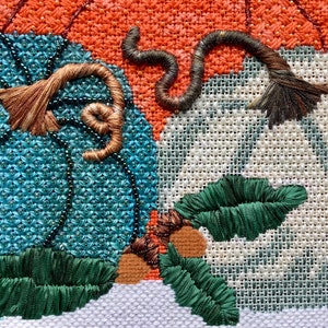 May include: Close-up of a cross-stitch embroidery featuring three pumpkins. The pumpkins are in shades of orange, teal, and white. The pumpkins have brown stems and green leaves.