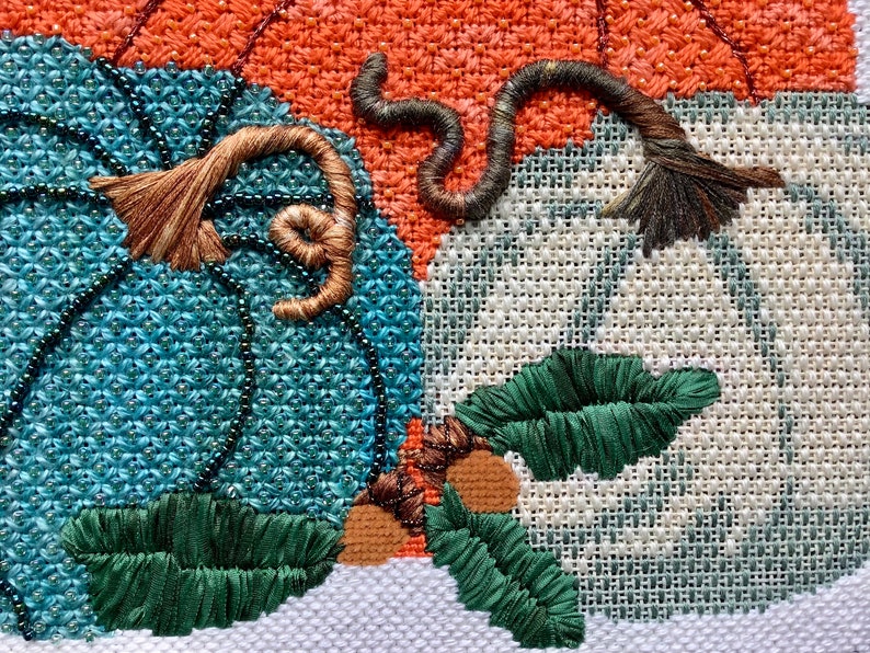 May include: Close-up of a cross-stitch embroidery featuring three pumpkins. The pumpkins are in shades of orange, teal, and white. The pumpkins have brown stems and green leaves.
