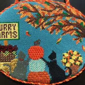 May include: A needlepoint design featuring two cats sitting under a tree with fall leaves. The sign reads "Purry Farms".