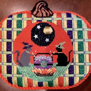 May include: A hand-stitched pumpkin-shaped pillow with a plaid background and a Halloween scene featuring two black cats, a cauldron, and a crescent moon with stars.