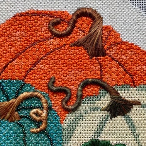 May include: Close-up of a hand-stitched embroidery design featuring three pumpkins. The pumpkins are orange, teal, and white with brown stems. The stems are curled and have a frayed texture.