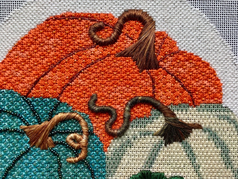 May include: Close-up of a hand-stitched embroidery design featuring three pumpkins. The pumpkins are orange, teal, and white with brown stems. The stems are curled and have a frayed texture.