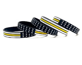 May include: Four black silicone bracelets with a thin yellow stripe and white stars. The bracelets are stacked on top of each other.