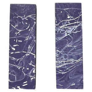 May include: Two rectangular bookends with a marbled design. The bookends are primarily a deep purple color with white streaks and splashes throughout. The design gives the bookends a unique, artistic look.
