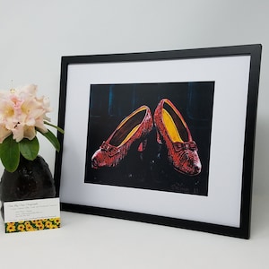 May include: A framed print of a painting depicting a pair of red shoes with a black background. The shoes are a popular symbol from the movie "The Wizard of Oz".